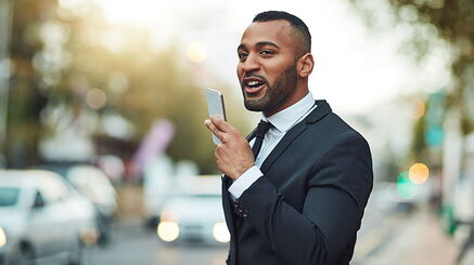 Ein junger Mann im Anzug hält sich ein Smartphone vor das Gesicht, in das er hinein spricht.